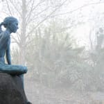 A bronze statue of a girl sitting on a large boulder, set in an outdoor garden. She gazes contemplatively at her surroundings, with her hair tied back, blending naturally with the misty greenery and shrubs in the background.