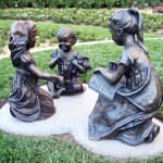 Bronze sculptures of three young girls seated in a circle on a grass-covered garden pathway. One girl holds an open book, another clutches a teddy bear, and the third engages attentively, creating a serene and playful storytelling scene. The setting features vibrant rose bushes and a tranquil garden backdrop.