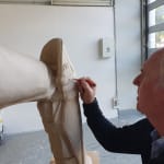 Allan Border signs his initials on the clay model of his statue during the sculpting process in Linda Klarfeld's studio. The close-up highlights the artist's attention to detail in crafting the cricket pads and uniform.