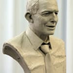 An angled view of A clay bust sculpture of Anthony John Abbott, former Prime Minister of Australia, in progress. The bust features detailed facial expressions and a textured surface, showing Abbott in a collared shirt and tie. The unfinished texture highlights the sculpting process, emphasizing the artist's craftsmanship. The background is neutral with soft, white draping, drawing attention to the clay model.