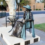 A close side view of the bronze statue of Mayor Joy Baluch, focusing on her poised and elegant posture as she sits in a chair next to a table. The background shows urban surroundings with a tree and public structures.