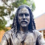 A close-up of the bronze statue depicting Jesus with detailed facial features, including flowing hair and a beard, set against a natural background with trees in soft focus.