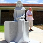 A granite statue of a seated woman in a flowing gown holding a book, with a man in casual clothing standing nearby under a bright blue sky.