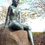 A life-size bronze sculpture of a young girl sitting on a smooth boulder outdoors, surrounded by golden autumn leaves. The girl’s playful expression and relaxed posture exude a sense of peace and harmony with the natural environment.