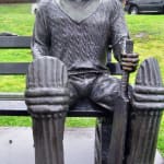 Bronze statue of Bill Woodfull, a legendary cricketer, seated on a bench holding a cricket bat, with protective gear on his legs. The detailed expression and attire honor his contributions to the sport.