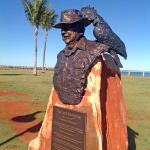 Angled view of the bronze bust of Lord Alistair McAlpine mounted on a rust-colored stone pedestal, with an engraved plaque below. The sculpture features McAlpine wearing a wide-brimmed hat and a cockatoo perched on its brim. The statue is set in an open grassy area with a clear blue sky and ocean visible in the background.