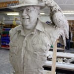 Angled Close-up of a clay bust of Lord Alistair McAlpine in progress, wearing a wide-brimmed hat and a textured button-up shirt. A detailed black cockatoo perches on the brim of the hat, adding a unique element to the sculpture. The bust captures a warm and smiling expression