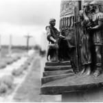 A bronze relief sculpture depicting Station I of the Stations of the Cross, titled 'Jesus Before Pilate.' The artwork shows Pilate seated on a throne-like chair, gesturing towards Jesus, who stands bound and flanked by Roman soldiers. The detailed architectural backdrop features columns and a staircase, symbolizing the Roman authority. The foreground provides a sense of depth, while the distant pathway hints at the larger Stations of the Cross installation.