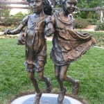Bronze sculpture of two young girls holding hands and skipping, capturing movement and joy, part of the Expressions of Love series.