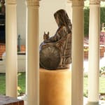 Rear view of the bronze statue of St. Mary MacKillop and her dog, Bobbs, seated in a wheelchair. The statue is surrounded by decorative columns and set on a pedestal, highlighting her enduring spirit and humility.