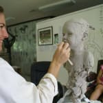 Artist sculpting a clay portrait bust of a young child in a studio, with the child model seated in the background wearing a colorful dress and smiling. The scene highlights the creative process and attention to detail in capturing the child's likeness.