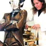 The sculptor, Linda Klarfeld, adjusts the stacked gold bullion on the backrest of the large banker sculpture. The seated figure, dressed in a formal suit with a reflective polished face, embodies wealth and power with its striking design.