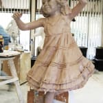Clay sculpture of a joyful young girl in a ruffled dress, captured mid-dance with an arm raised gracefully, displayed in a studio setting with sculpting materials and other works visible in the background.