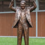 A bronze statue of Nick Columb standing on a green lawn, with both arms raised in a victorious gesture. He is depicted in a suit and tie, with an exuberant expression, holding a book in one hand. The statue is set outdoors, with a wooden stable-like structure in the background, symbolizing his achievements in the horse racing industry.