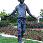 Bronze sculpture of a young boy balancing on a log, part of a whimsical children's garden scene in a lush park setting.