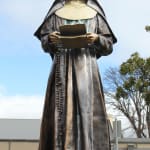 Full view of the bronze statue of St. Mary MacKillop set against a backdrop of historic church architecture. The statue, holding a letter from Fr Julian, symbolizes her contributions the faith.