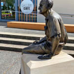 A side profile view of the completed bronze statue of Max Watters. The statue is positioned outside the Muswellbrook Regional Arts Centre, with a sign for the "Gallery Café" visible in the background.