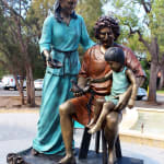 A full view of the bronze statue of the Holy Family placed outdoors. Mary, Joseph, and the child Jesus are depicted in a peaceful family interaction. Joseph is seated, holding the child on his lap with a dove in his hands, while Mary stands beside them, her hand resting on Joseph’s shoulder. The base of the statue is inscribed with the words "Seek Truth." The background features trees and a sunny sky.