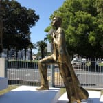 A bronze statue of a young man stepping forward confidently, draped in a cloak resembling an Australian flag. The sculpture is mounted on a white platform with engraved text and dates, set outdoors in a landscaped area with trees, a fence, and a street in the background under a clear blue sky.
