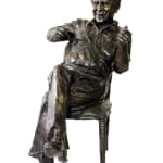 Bronze statuette depicting Norman, seated casually on a chair. He has a cheerful expression with a wide smile, his legs crossed, and his hands gesturing animatedly, capturing a lively and relaxed demeanor. The textured details of the sculpture highlight his clothing and posture.