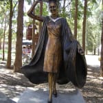 A bronze statue of a female graduate at Macquarie University, depicted joyfully holding her mortarboard aloft in celebration. She wears academic robes that flow dramatically behind her, symbolizing achievement and pride.