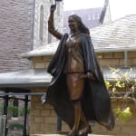 A bronze statue of a young woman dressed in academic regalia, including a gown and mortarboard, celebrating her graduation. She holds a diploma in her left hand and raises her cap high with her right hand. Her face is lit with a joyful smile. The statue is placed on a sandstone pedestal in front of the Manly International School of Management, with the historic stone building in the background.