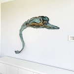A bronze relief sculpture titled "Freestyle" mounted on a white wall. The artwork depicts a swimmer gliding forward with fluid movement, highlighted by textured waves and a flowing form. The surrounding area is minimal, featuring a sleek white cabinet below the sculpture.