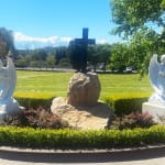 Two marble angel statues flank a large stone cross in a landscaped setting. The scene, set amidst a well-manicured garden, reflects a peaceful memorial atmosphere under a sunny sky.