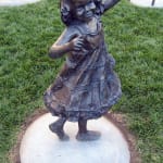 Bronze sculpture of a joyful young girl in a ruffled dress, captured mid-dance with an arm raised gracefully, In a garden setting with other bronze statues of children visible in the background.