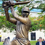 Detail of The bronze statue of Allan Border at The Gabba in Brisbane depicts the cricketer mid-swing in an iconic pose. The intricate details of the bat, pads, and uniform celebrate his legacy. The statue is mounted on a sandstone base with a commemorative plaque, surrounded by greenery and people at the unveiling event.