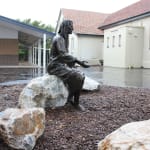 Angled side view of a bronze statue of Jesus seated on a rock during a rainy day, with a modern building in the background.