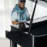 Bronze sculpture of an elderly man seated at a grand piano, playing with a serene and focused expression. The figure is dressed casually in shorts, a t-shirt, and a sweater draped over his shoulders, with vibrant color accents highlighting his clothing and shoes. The artwork captures a moment of musical engagement and nostalgia in a modern indoor setting.