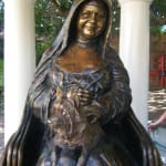Close-up of a bronze statue of St. Mary MacKillop seated with a gentle smile, holding her beloved dog, Bobbs, in her lap. The finely detailed sculpture captures her warmth and compassion.