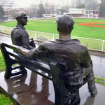 Bronze sculptures of cricketers Bill Woodfull and Keith Miller seated on a bench, overlooking a cricket field. The figures, depicted in casual poses, evoke a sense of camaraderie and nostalgia in a rainy outdoor setting.