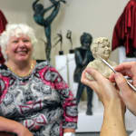 A close-up photograph of Linda Klarfeld sculpting a mini-bust of Marina, a smiling woman seated in the background. Marina, wearing a red and black patterned outfit and a gold chain necklace, is cheerfully observing the sculpting process. The detailed bust in progress captures her likeness with impressive accuracy, set in a studio adorned with other sculptures and artworks in the background.