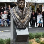 A bronze bust of Dom Lopez, sculpted by Linda Klarfeld, displayed outdoors during its unveiling ceremony in Mosman. The bust portrays Dom Lopez wearing the ceremonial mayoral chain, showcasing intricate details of the chain's design. It sits atop a granite pedestal inscribed with Dom Lopez’s name, title, and a commemorative plaque detailing his life and contributions to the Mosman community. Attendees of the ceremony, seated in the background, watch as the sculpture is revealed.