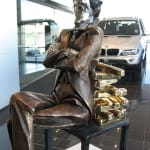 The large banker sculpture showcased in a car dealership, with luxury vehicles in the background. The figure, seated on a chair with gold bullion forming the backrest, conveys themes of wealth and affluence. The polished reflective face adds a modern, surreal touch to the piece.