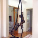 Side view of a bronze sculpture of two young boys playing on ropes, displayed indoors on a polished wooden surface. One boy climbs the rope confidently while the other, slightly younger, joyfully swings from a looped section, their expressions capturing a playful and adventurous spirit. Behind the sculpture is a bookshelf filled with framed family photographs, adding a warm, personal backdrop to the scene.