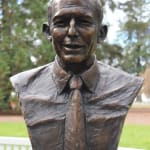 A bronze bust sculpture of Anthony John Abbott, former Prime Minister of Australia in a shirt and tie, set against an outdoor backdrop of lush green trees. The sculpture captures the man's detailed facial features, including a slight smile, closely cropped hair, and defined cheekbones. The textured surface of the bronze gives the piece a dynamic and expressive quality, emphasizing the character and strength of the subject. The bust is mounted on a granite pedestal.
