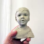 A small clay bust of a young boy with short hair, sculpted in plasticine. The figure has a soft, expressive face with slightly parted lips, capturing a sense of innocence. The bust is held in someone's hand against a plain white background.