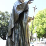A bronze statue of Pope John Paul II depicted in a blessing gesture, with one hand raised and the other holding a staff topped with a crucifix. The statue features flowing robes and a detailed mitre. It is set outdoors, with trees and a fence visible in the background. A purple flower lies at the base of the statue.