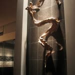 The Tinkerbell sculpture captured at night, illuminated by warm lighting that emphasizes the intricate details of the bronze and its dramatic shadows on the wall. The figure's fluid lines and sweeping forms create an ethereal effect.