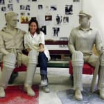 The artist seated between clay models of cricketers Keith Miller and Bill Woodfull during the sculpting process. The unfinished statues showcase early stages of detailing and craftsmanship in a studio filled with reference photos.