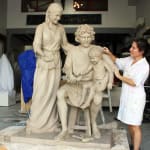 A clay sculpture of the Holy Family in progress, featuring Mary, Joseph, and the child Jesus. Joseph is seated on a stool, holding the child on his lap, guiding his hands toward a dove. Mary stands beside them, gently touching Joseph's shoulder, with a soft, caring expression. Linda Klarfeld the artist is seen working on the sculpture, adding details to Joseph’s shoulder.