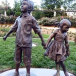 ronze sculpture of a boy and girl standing side by side, holding hands. The boy looks ahead while the girl glances down, showcasing their innocence and connection.