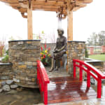 A bronze statue of Mrs. Jen seated on a stone bench under a wooden pavilion with intricate beams. The statue depicts her reading a book, exuding a sense of tranquility. A vibrant red wooden bridge leads to the pavilion, surrounded by cobblestone paths and stone walls adorned with colorful flowers. The setting is serene, despite the wet weather creating a reflective sheen on the surfaces.