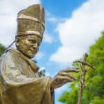 Detail of a bronze sculpture of St John Paul II holding a staff topped with a crucifix.. The scene is set outdoors on a sunny day, with a tree in the background.