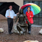 A bronze statue of Jesus seated on a rock with Fr Peter and sculptor Linda Klarfeld standing beside it, holding umbrellas, one wearing a red coat and holding a colorful rainbow umbrella.