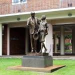 A full view of the completed Shore Boys bronze statue, installed at Shore Grammar School. The sculpture portrays a man guiding a young boy in school uniform, holding a hat and books. The base features the engraved word "LAMPADA," symbolizing passing the torch.