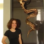 A bronze sculpture titled Tinkerbell by Linda Klarfeld mounted on a glossy black tiled wall. The sculpture depicts a dynamic female figure with flowing forms that resemble wings and a sweeping tail, evoking a sense of motion. The artist, Linda Klarfeld, stands smiling beside the artwork, wearing a black shirt and jeans.